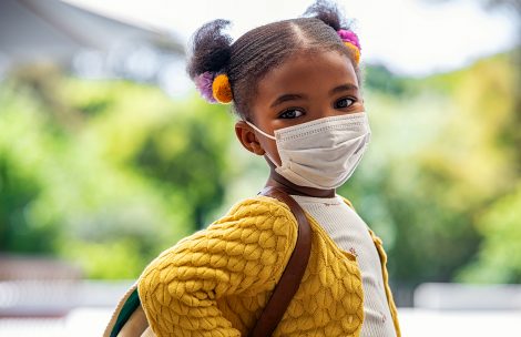 Smiling cute little girl with school backpack and protective face mask ready for first day of school during covid pandemic. African american female child wearing surgical mask while looking at camera. Cute black kid going back to school during coronavirus pandemic disease.