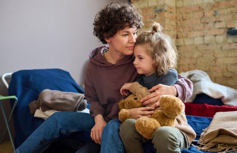 Young woman giving hug to her child with brown soft Teddy bear while both are sitting on a bed.