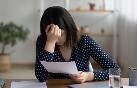 Close up frustrated Asian woman reading bad news in letter, sitting at work table, upset depressed businesswoman covering face, shocked by fraudulent letter.
