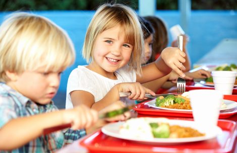 Elementary Pupils Enjoying Healthy Lunch In Cafeteria Sitting Down Smiling At Camera