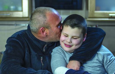 Dad and his son with autism sitting in kitchen at home. A man with short hair and bristle hugs his son and kisses him on the cheek.