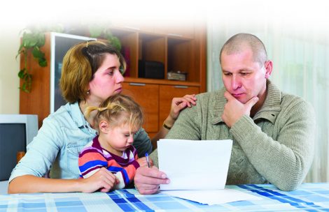 A mother and father look worried as they assess bills and financial papers. Their child sits at a table with them.